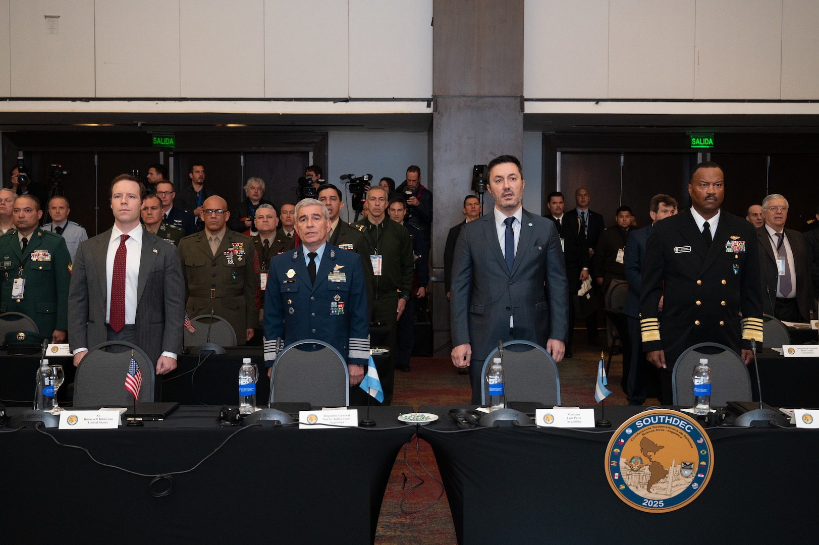 Two men in grey suits and two men in military uniforms stand in a line behind a table