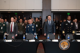 Two men in grey suits and two men in military uniforms stand in a line behind a table