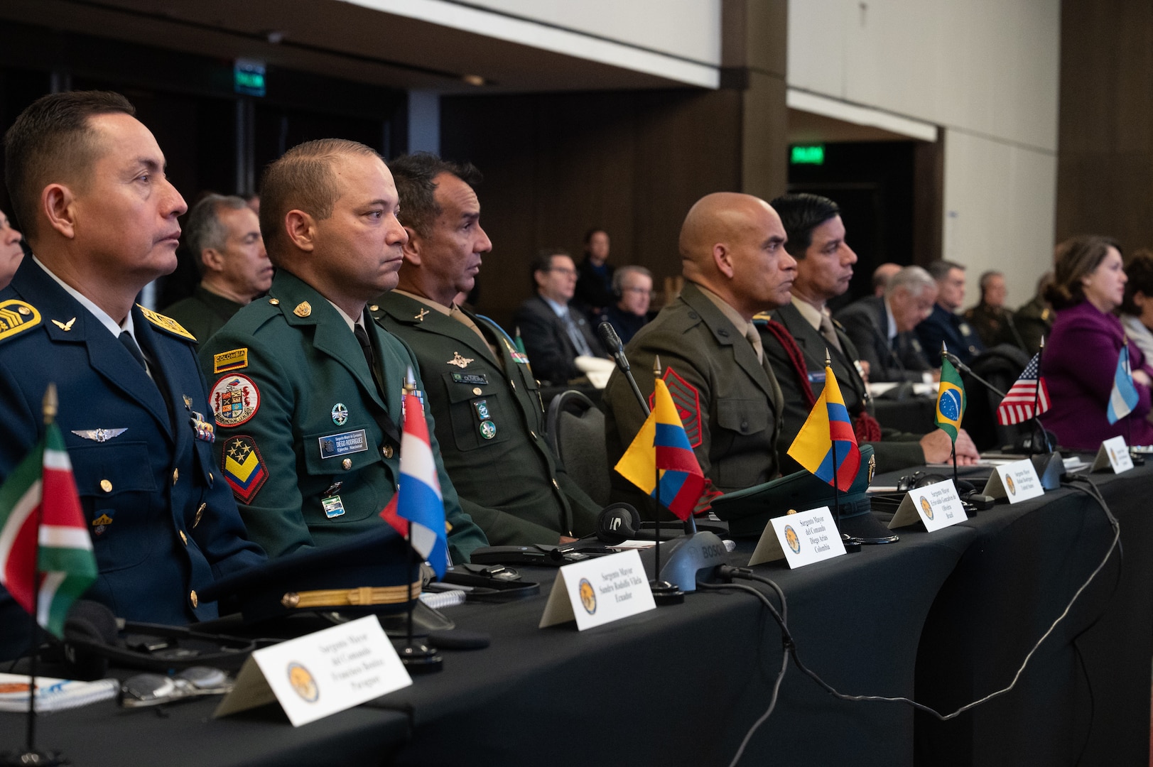A line of men in various military uniforms sit at a long table and listen to remarks.