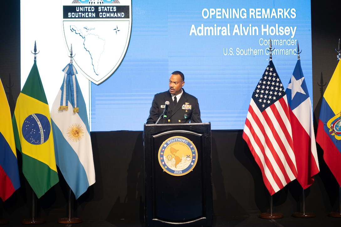 A man in a black military uniform speaks at a podium flanked on each side by various nation flags