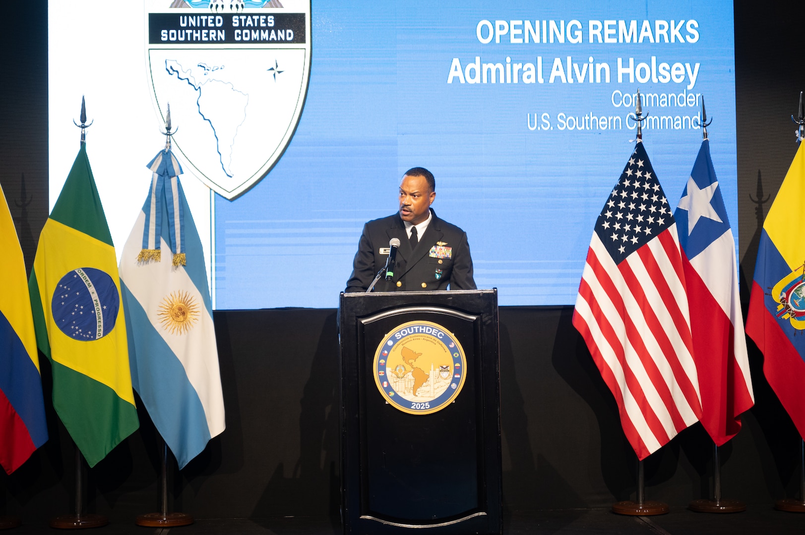 A man in a black military uniform speaks at a podium flanked on each side by various nation flags