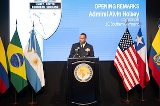A man in a black military uniform speaks at a podium flanked on each side by various nation flags