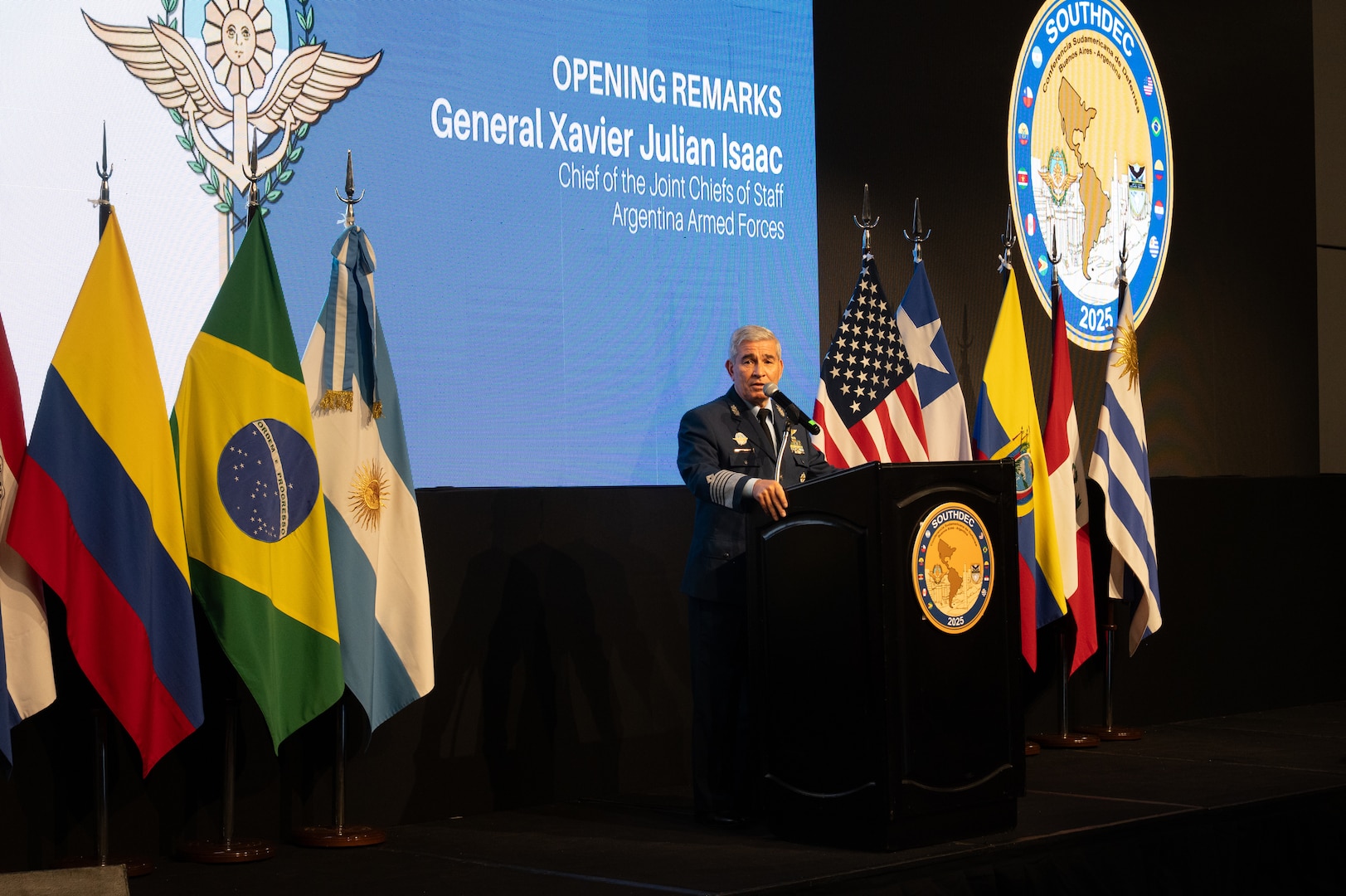 A man in a blue military uniform speaks at a podium flanked on each side by various nation flags.