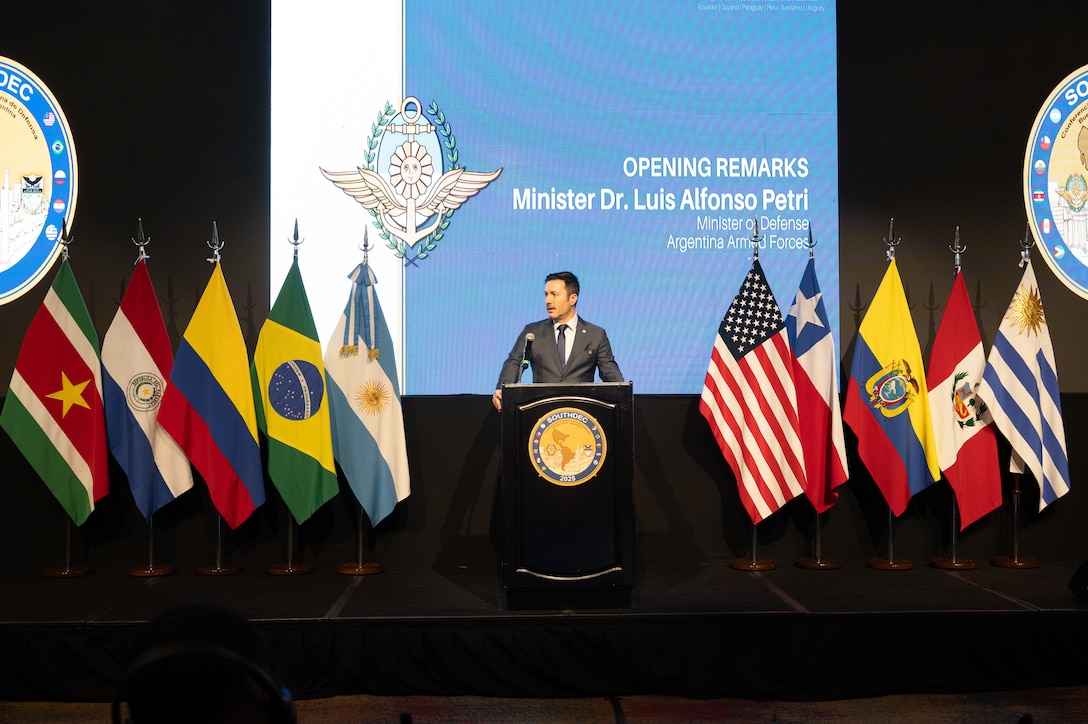 A man in a grey suit speaks at a podium flanked on each side by various nation flags.