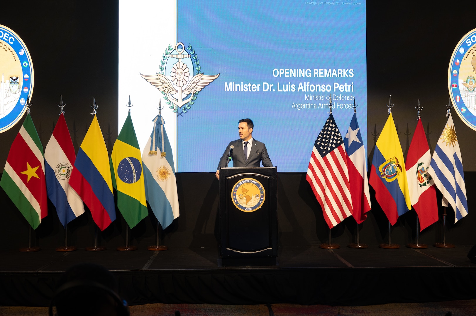 A man in a grey suit speaks at a podium flanked on each side by various nation flags.