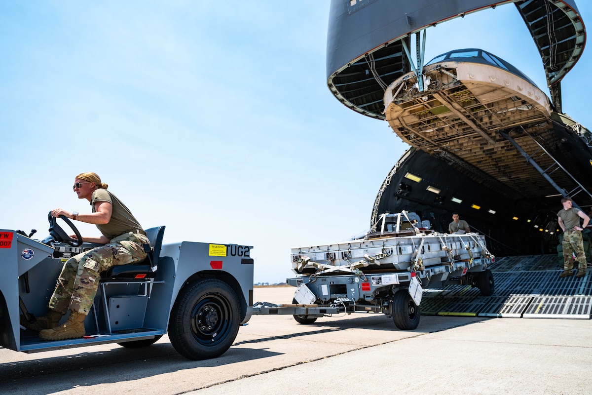 An airman drives a vehicle with an attached trailer loaded with cargo away from the open cargo bay of an aircraft, as other airmen watch from the aircraft.