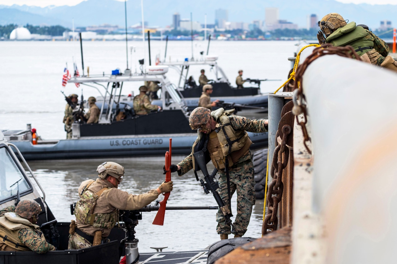 A service member on a boat hands a red service weapon to another service member on a dock while others watch and two boats float in the harbor behind.