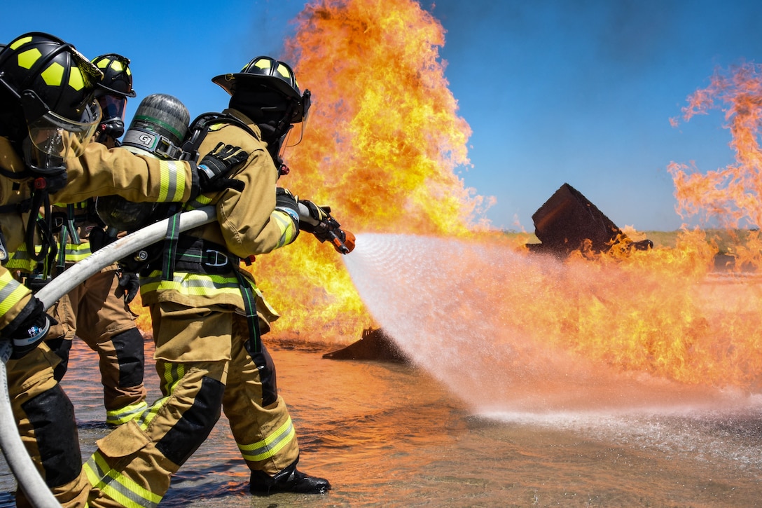 Three firefighters in gear hold a hose and spray flames outside under blue skies.