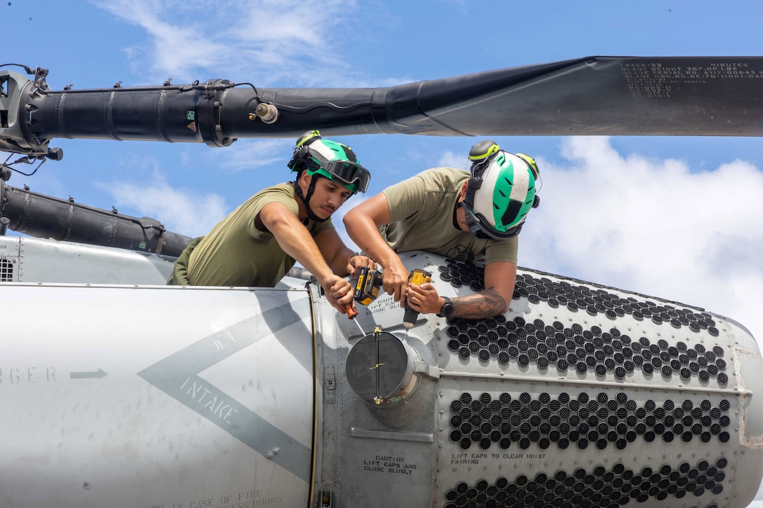 Two Marines in green and white helmets use tools on the side of a helicopter under blue skies.