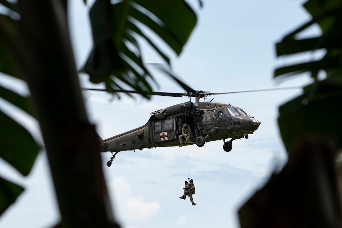 Two soldiers in a harness hang from a helicopter in flight against a cloudy sky as seen through a jungle environment.