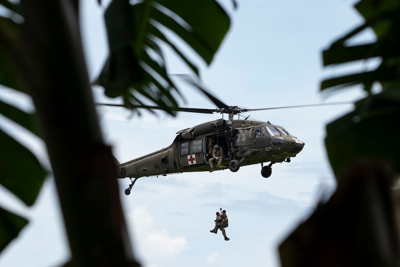 Two soldiers in a harness hang from a helicopter in flight against a cloudy sky as seen through a jungle environment.
