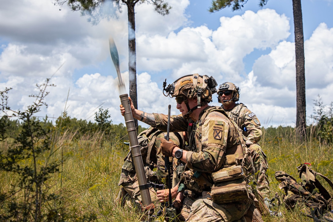 Soldiers in tactical gear kneel in tall grass and load a mortar into a weapon under partly cloudy skies with two trees behind.