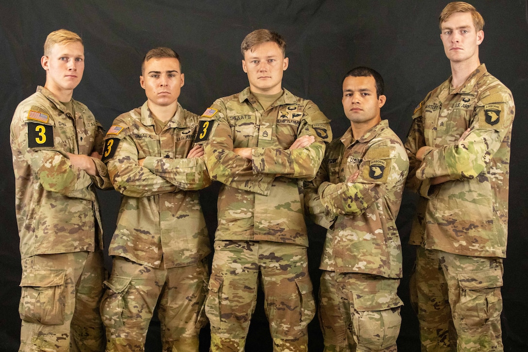 Five soldiers stand with their arms crossed against a dark backdrop.