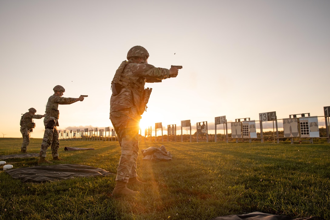 Three guardsmen in tactical gear stand and fire service weapons at targets in a field under a golden sky.