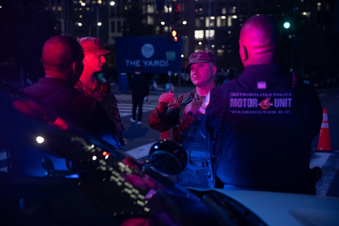 Two soldiers speak with two police officers who lean on a vehicle near a street illuminated with red light at night.