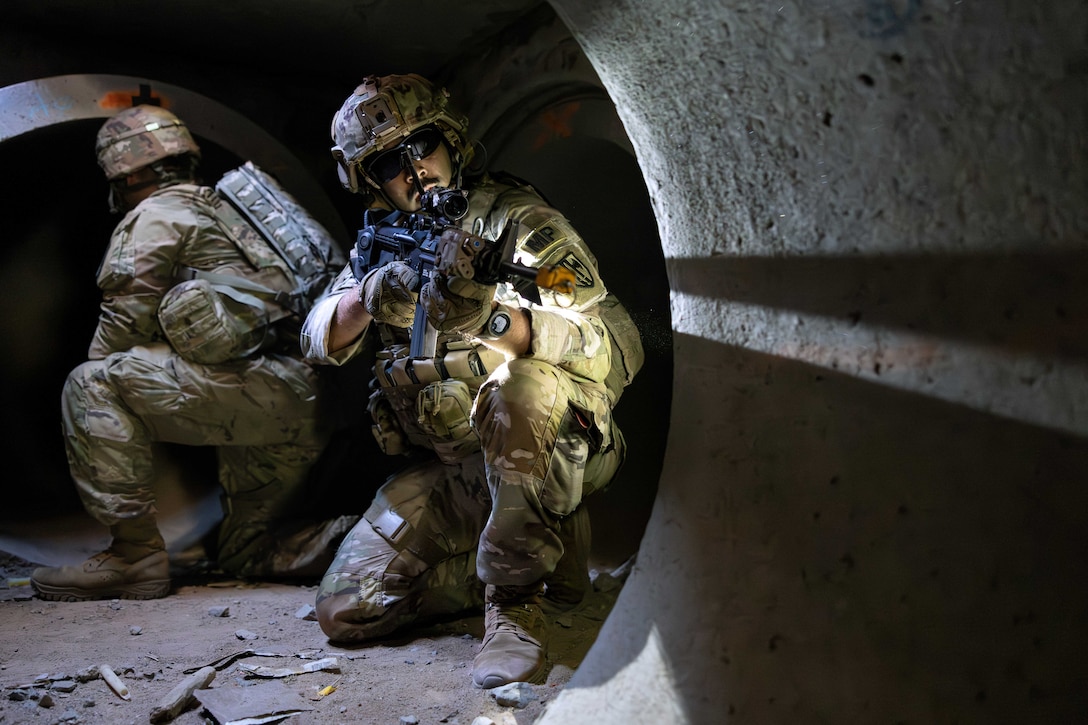 A soldier in tactical gear crouches in a tunnel and aims a service weapon as another soldier does the same while facing the other way.