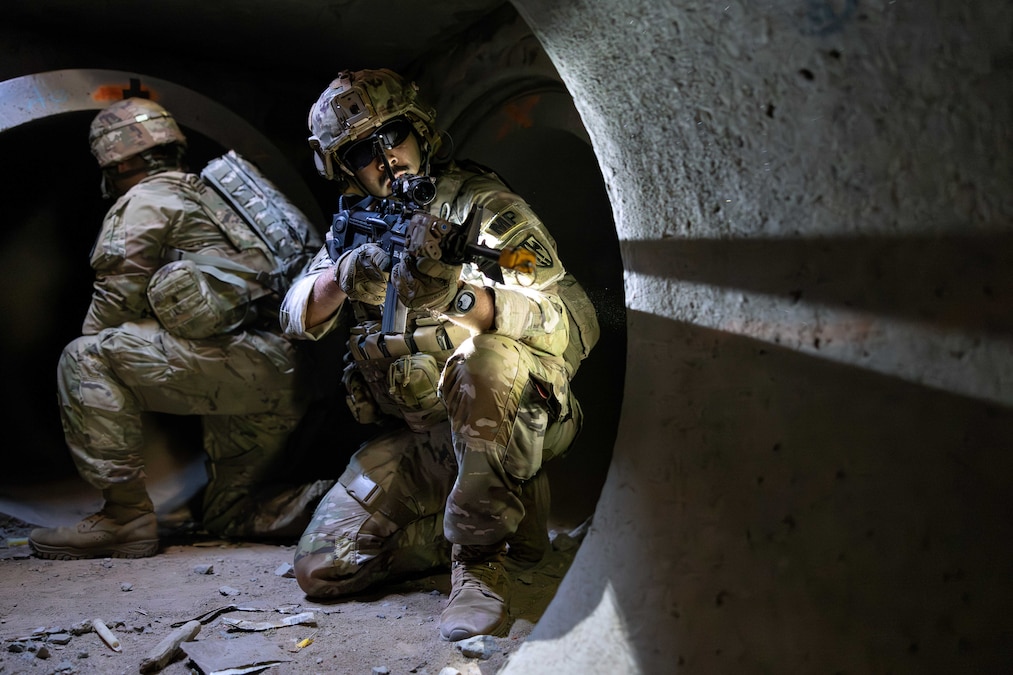 A soldier in tactical gear crouches in a tunnel and aims a service weapon as another soldier does the same while facing the other way.