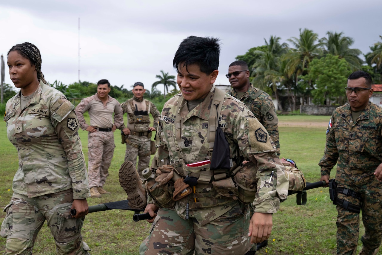 Four service members carry a stretcher with a simulated casualty as two more look on in the background on a grassy field during a cloudy day.