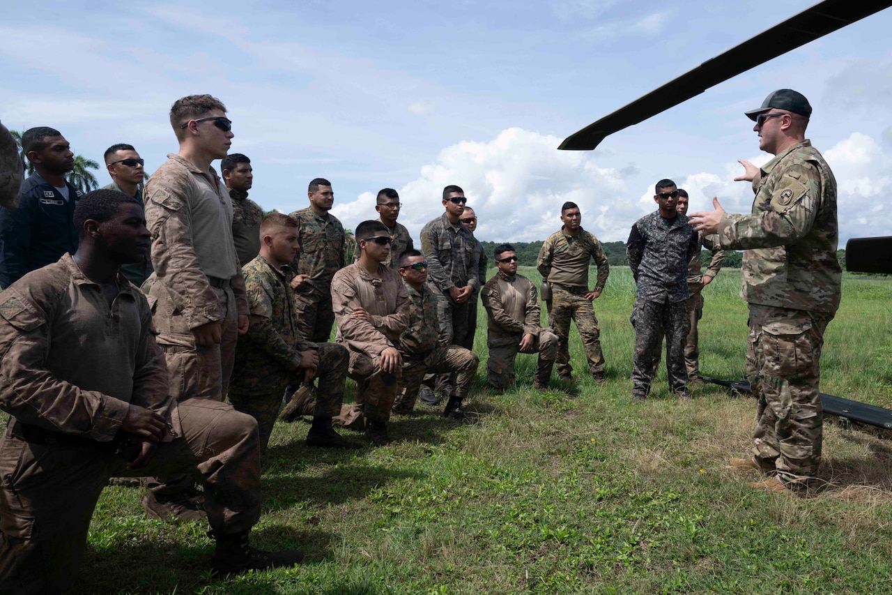 A military leader speaks to a group of multinational service members gathered on a grassy field outside during the day.