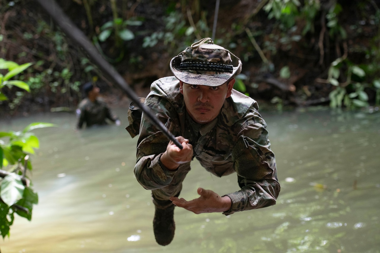 A Panamanian service member uses a rope suspended above water to cross a river in jungle.