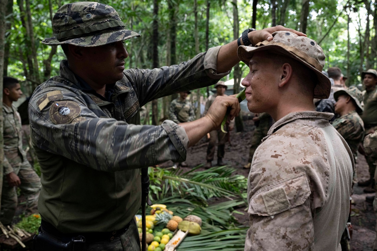 A Panamanian service member in uniform rubs a plant above the lips of a U.S. service member while outside in a jungle.