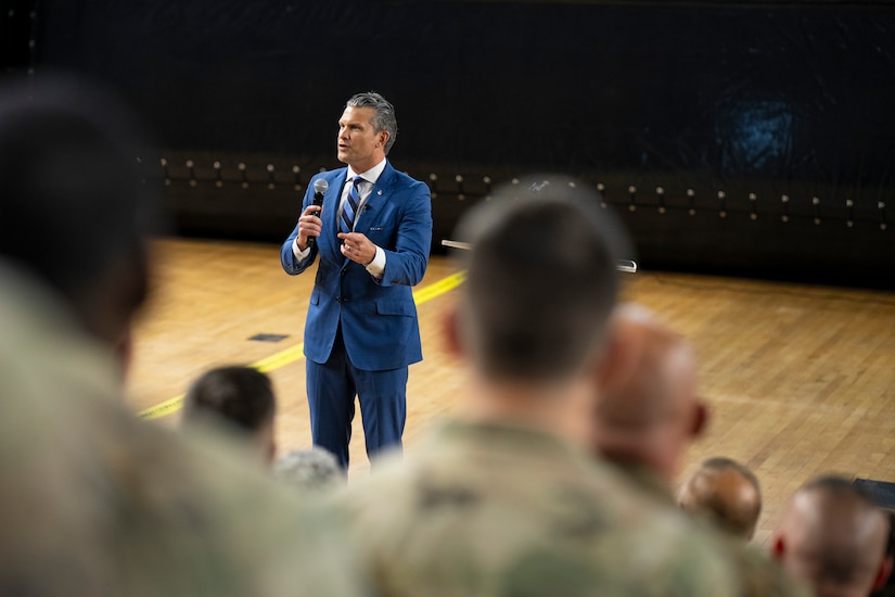 Defense Secretary Pete Hegseth holds and mic and speaks to a group of guardsmen in the stands.