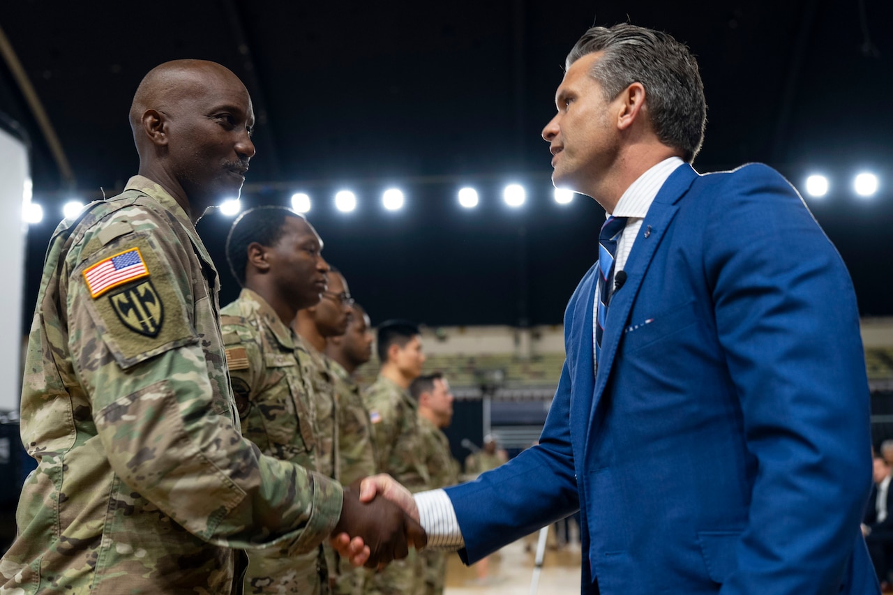 A person wearing a blue business suit shakes hands with a soldier as other soldiers stand in a line at an indoor facility.