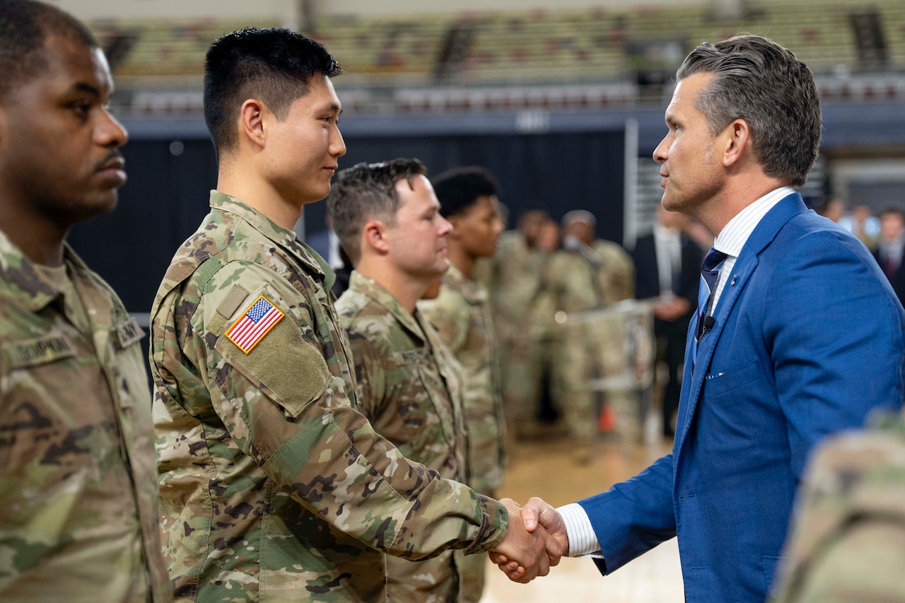 Defense Secretary Pete Hegseth shakes hands with one of three guardsmen standing in a line indoors with others in the background.