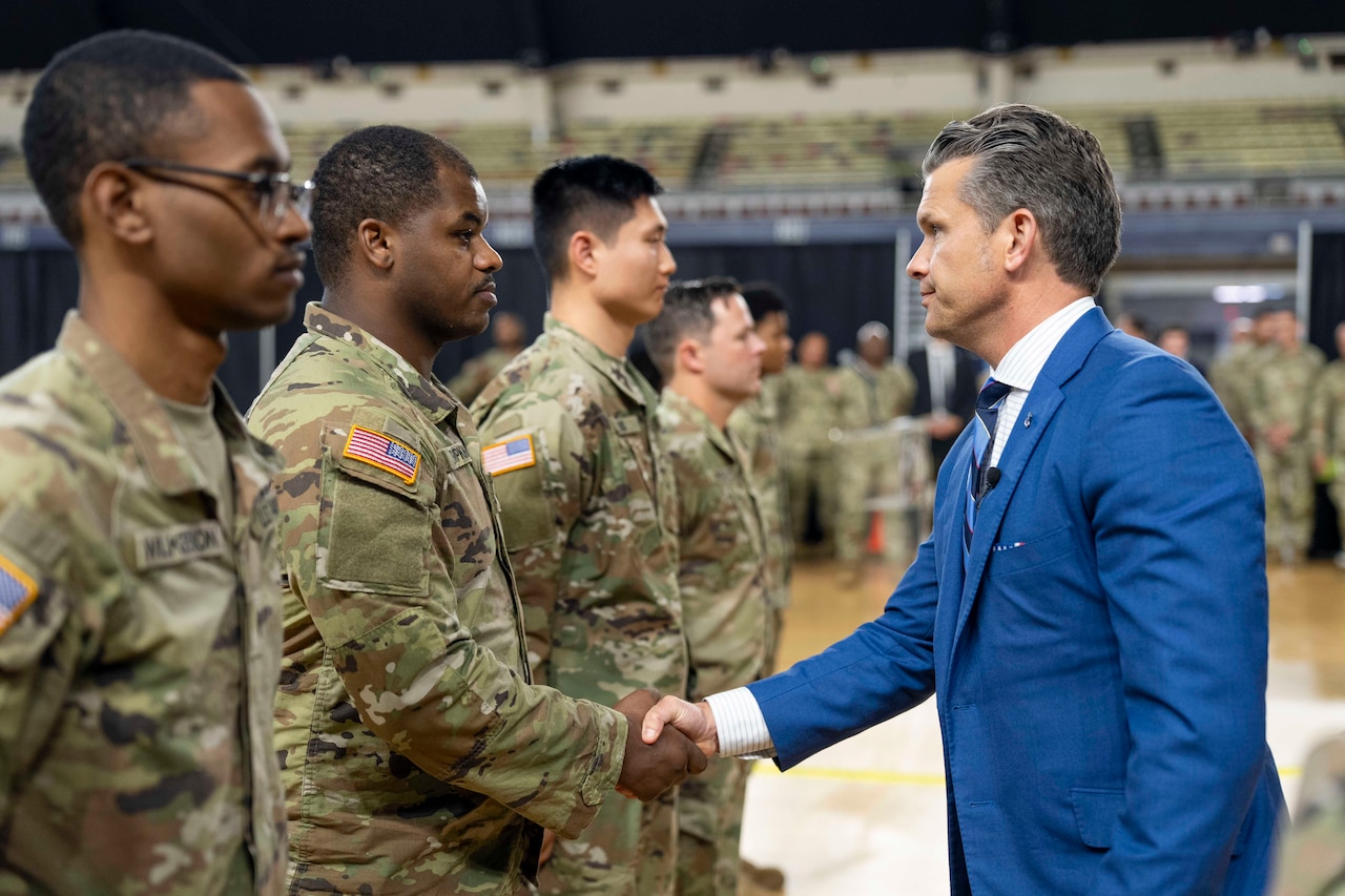 Defense Secretary Pete Hegseth shakes hands with one of several guardsmen standing in a line indoors with others in the background.