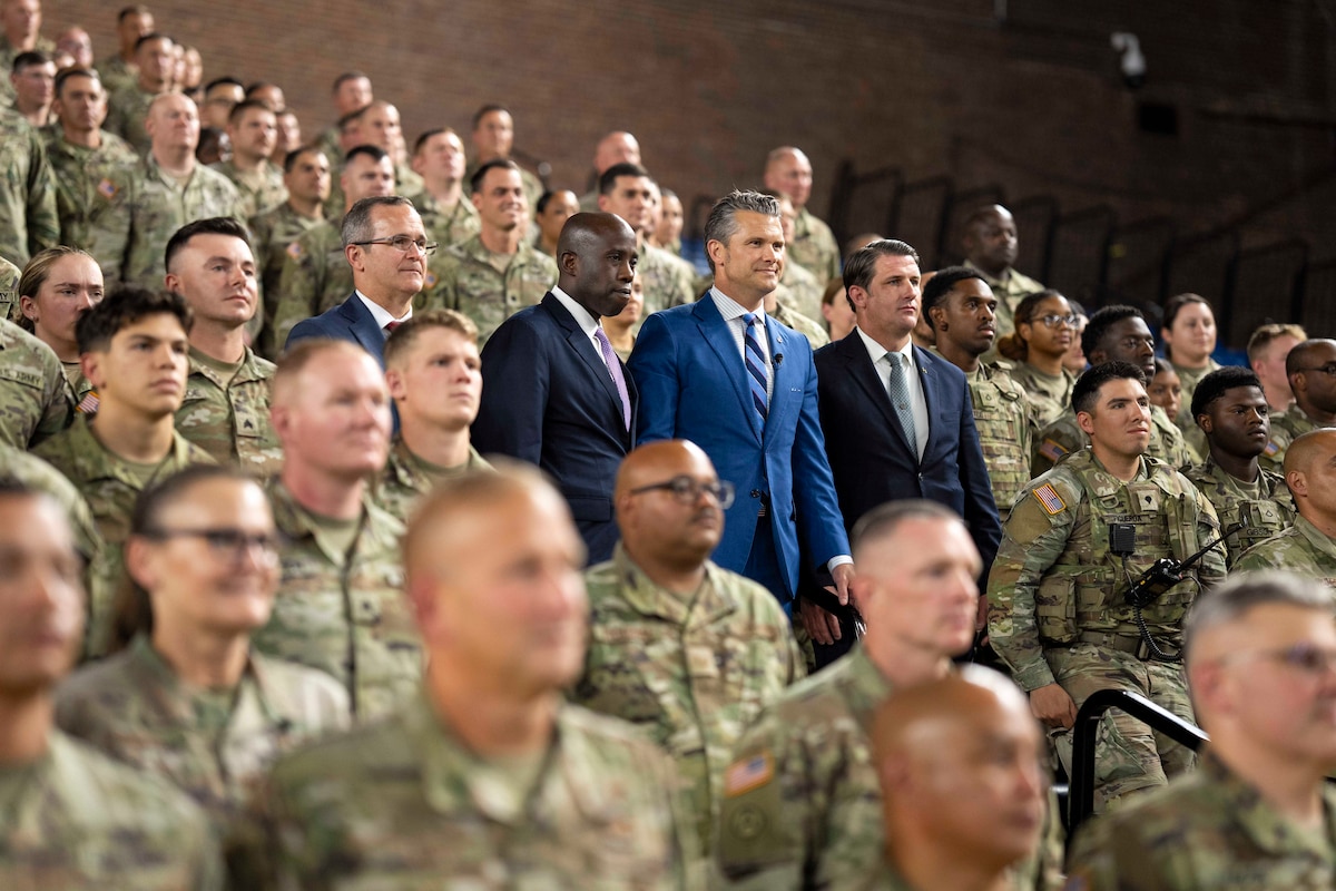 Defense Secretary Pete Hegseth and three other civilian leaders stand among a large group of National Guardsmen in a stadium-style room.