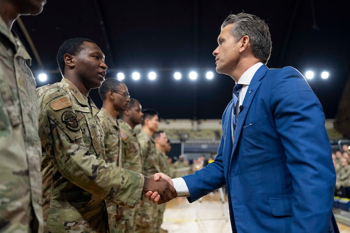 Defense Secretary Pete Hegseth shakes hands with one of several guardsmen in a line with a row of ceiling lights on in the background.