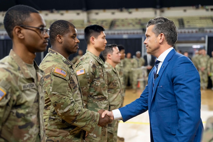 Defense Secretary Pete Hegseth shakes hands with one of several guardsmen standing in a line indoors with others in the background.