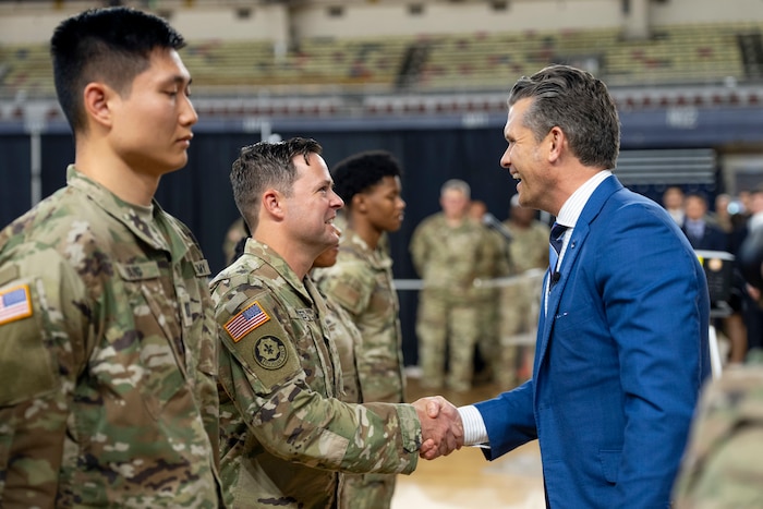 Defense Secretary Pete Hegseth laughs and shakes hands with one of several guardsmen in line, with others in the distance.