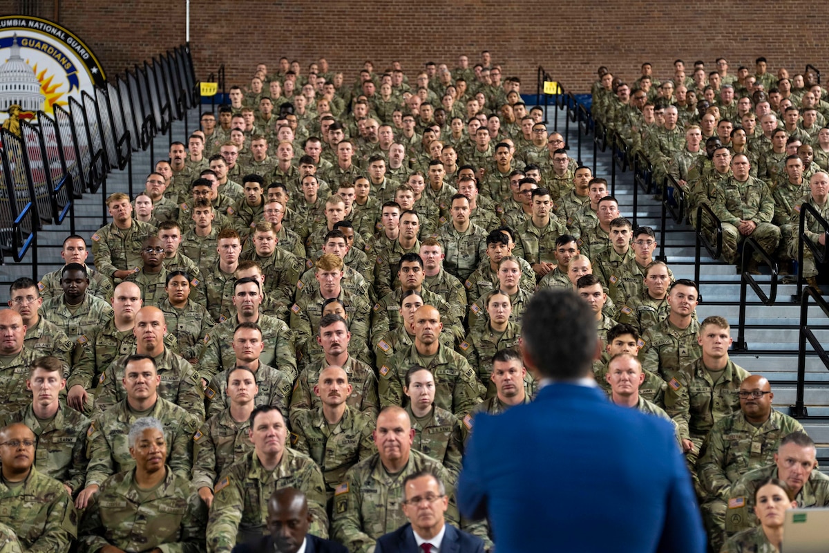 Defense Secretary Pete Hegseth is seen from behind as he speaks to a large group of guardsmen seated in stands in an auditorium.