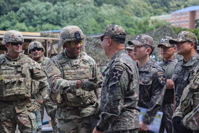 A U.S. and a South Korean service member shake hands outdoors as others look on.