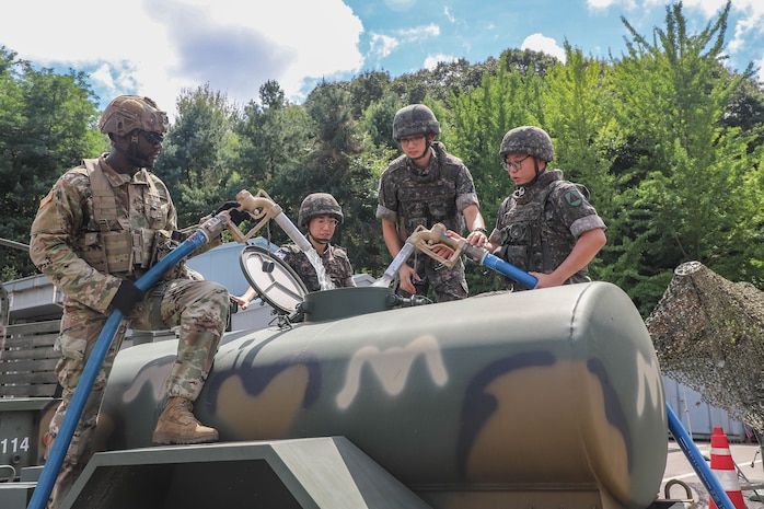 Two service members wearing military camouflage uniforms hold water hoses and fill up a water trailer as two others watch.