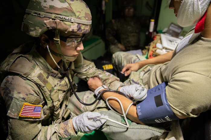 A soldier in uniform and ballistic helmet holds a stethoscope to a fellow soldier's arm while inflating a blood pressure cuff.