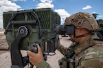 A man dressed in a military camouflage uniform and wearing a ballistic helmet looks through the viewfinder of a surveillance system in the desert.