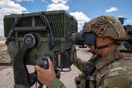 A man dressed in a military camouflage uniform and wearing a ballistic helmet looks through the viewfinder of a surveillance system in the desert.