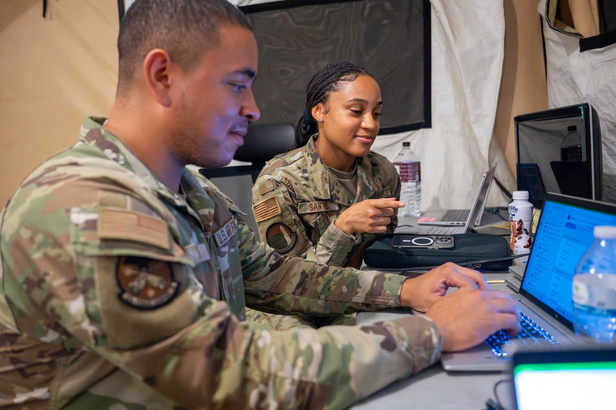 Two people look at a computer at a bare base encampment.