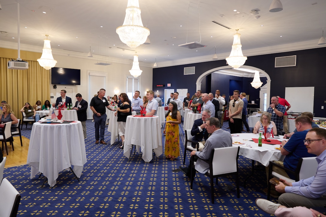 a gathering of people are scattered about a ballroom where white table cloth dressed round tables are adorned with napkins and centerpieces.