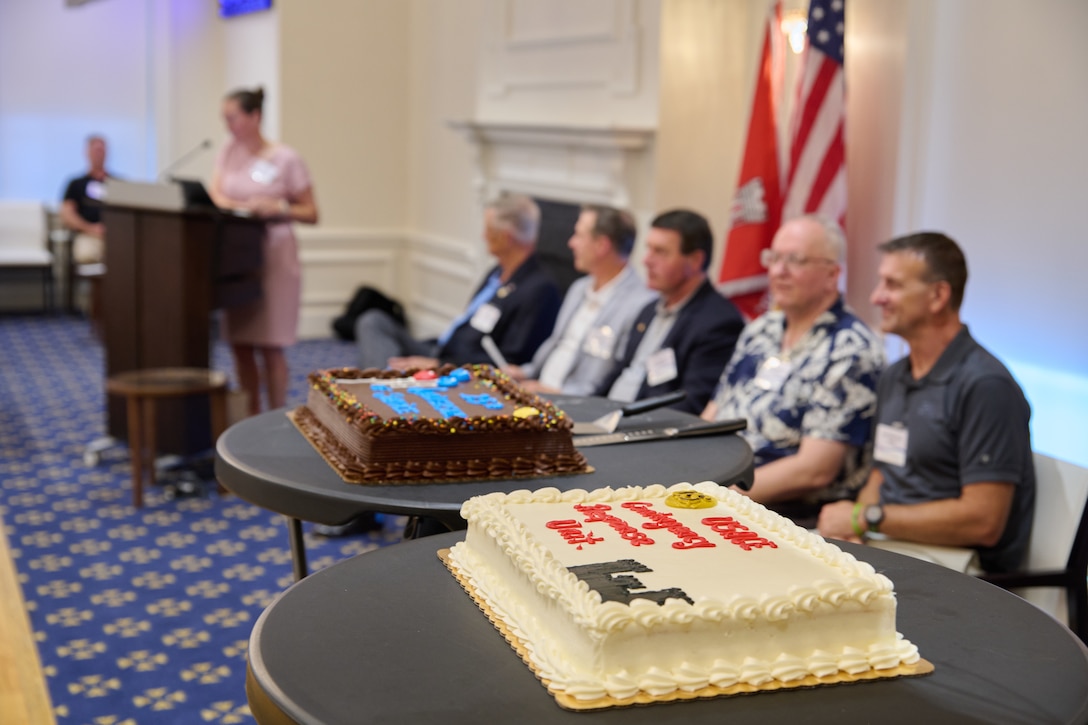 two cakes sit on tables behind them a group of men site while a woman speaks at a podium.