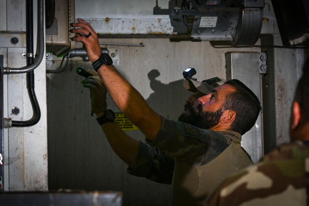 A person with a headlamp reaches up towards a panel.