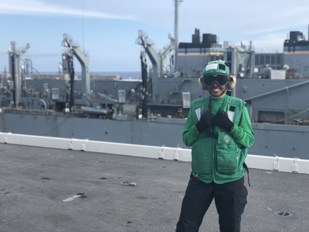 A woman wearing gloves, goggles, hearing protection, a green vest, green sweatshirt and black pants, smiles as she poses in front of a ship.