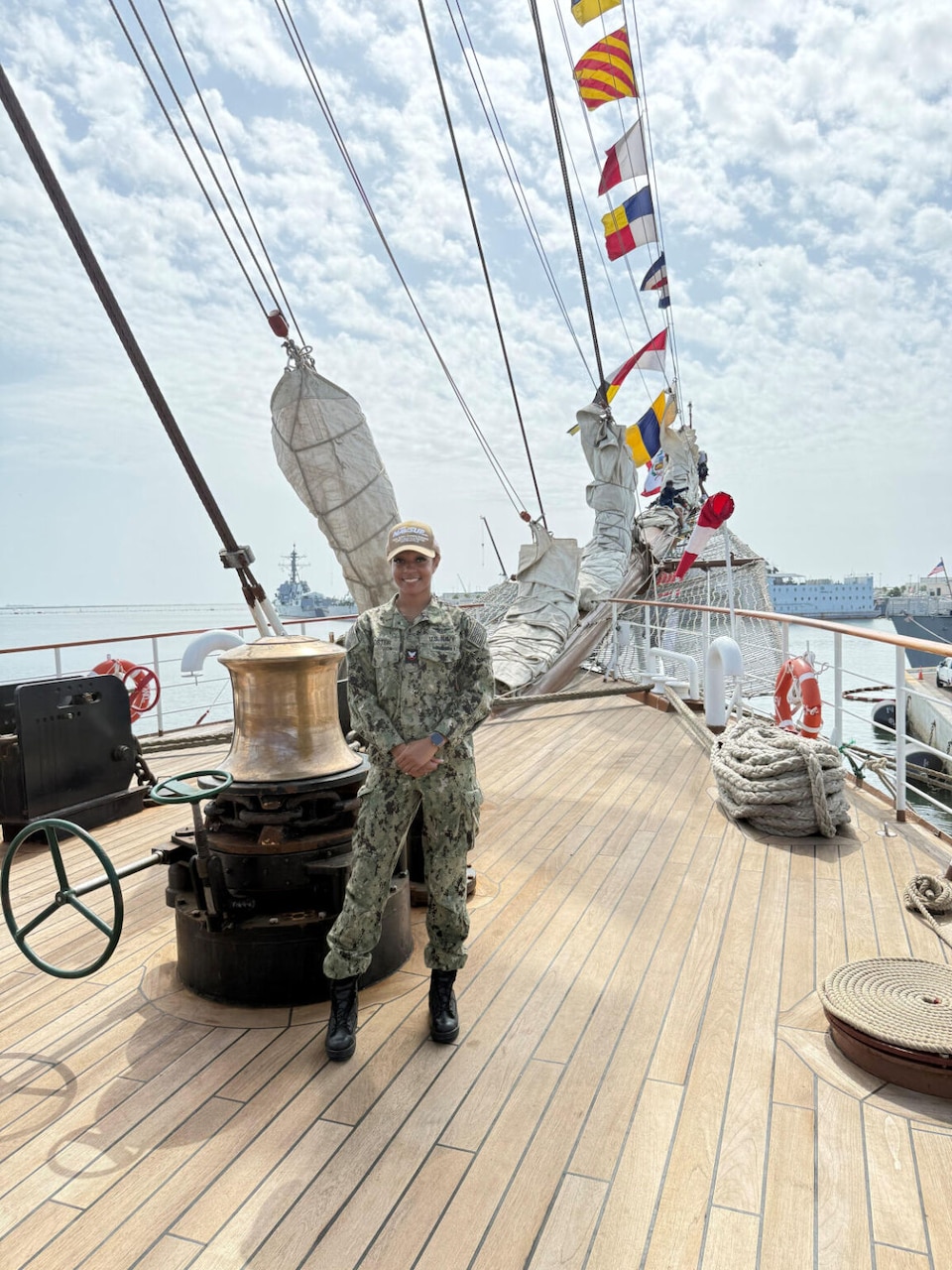 A woman wearing a military camouflage uniform and a tan baseball cap poses for a photo on the deck of a ship.