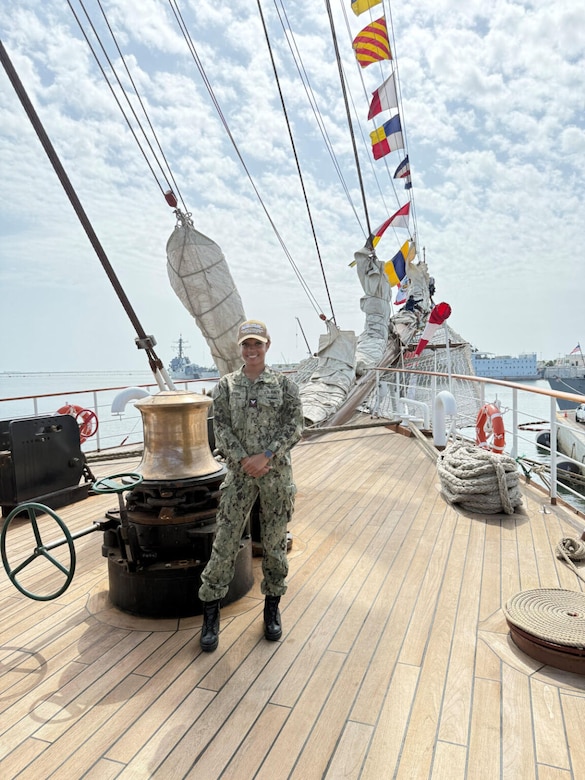 A woman wearing a military camouflage uniform and a tan baseball cap poses for a photo on the deck of a ship.