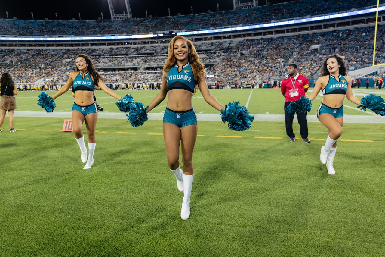 Three women dressed in shorts and crop top cheerleading outfits dance and wave pom poms held at their sides. There is a man wearing a red shirt and black slacks behind them. There is a stadium with people in the stands in the background.
