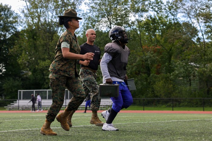 U.S. Marine Corps Sgt. Julie Leon, left, a drill instructor Golf Company, 2nd Recruit Training Battalion, Marine Corps Recruit Depot Parris Island, and a Los Angeles, California native, and Capt. Edgardo Diaz, center, the executive officer of Recruiting Station Boston, encourages a Randolph High School football player during the maneuver under fire portion of a Marine Corps combat fitness test as part of Marine Week Boston, Aug. 21, 2025. During Marine Week Boston, Marines with Marine Corps Recruiting Command shared Marine Corps traditions with local high schools in honor of the Corps’ 250th birthday. Marines stationed across the globe commemorate the 250th anniversary of the Marine Corps, honoring a distinguished legacy of service, sacrifice, and unwavering fidelity to both the nation and fellow Marines. (U.S. Marine Corps photo by Sgt. Aidan Hekker)