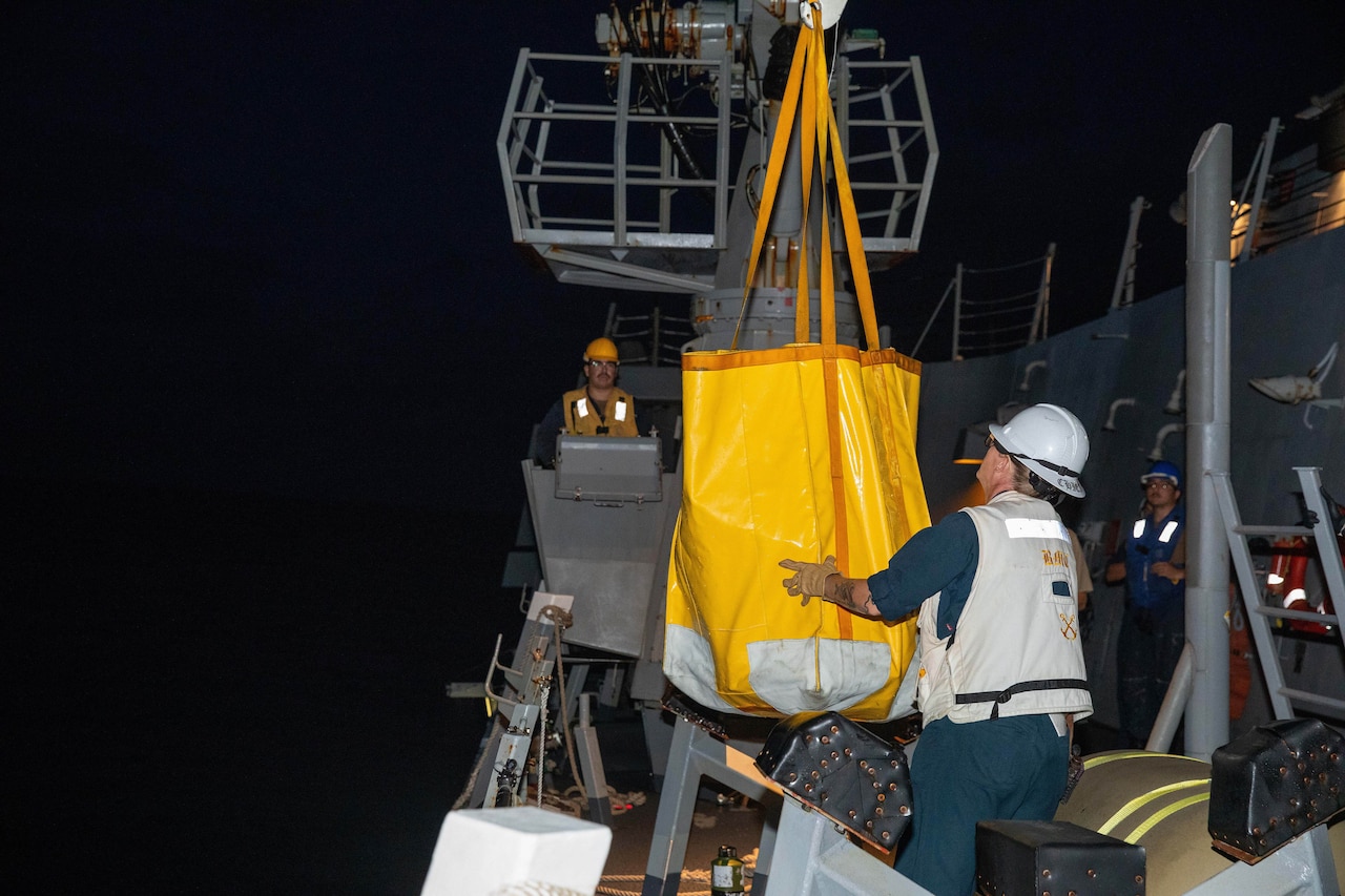 A sailor in a vest and safety helmet guides a large bag hanging from a crane, as a sailor in the background watches.