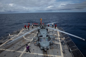 Sailors wearing red, white, green and purple long-sleeve T-shirts, walk around a helicopter on the deck of a ship at sea.